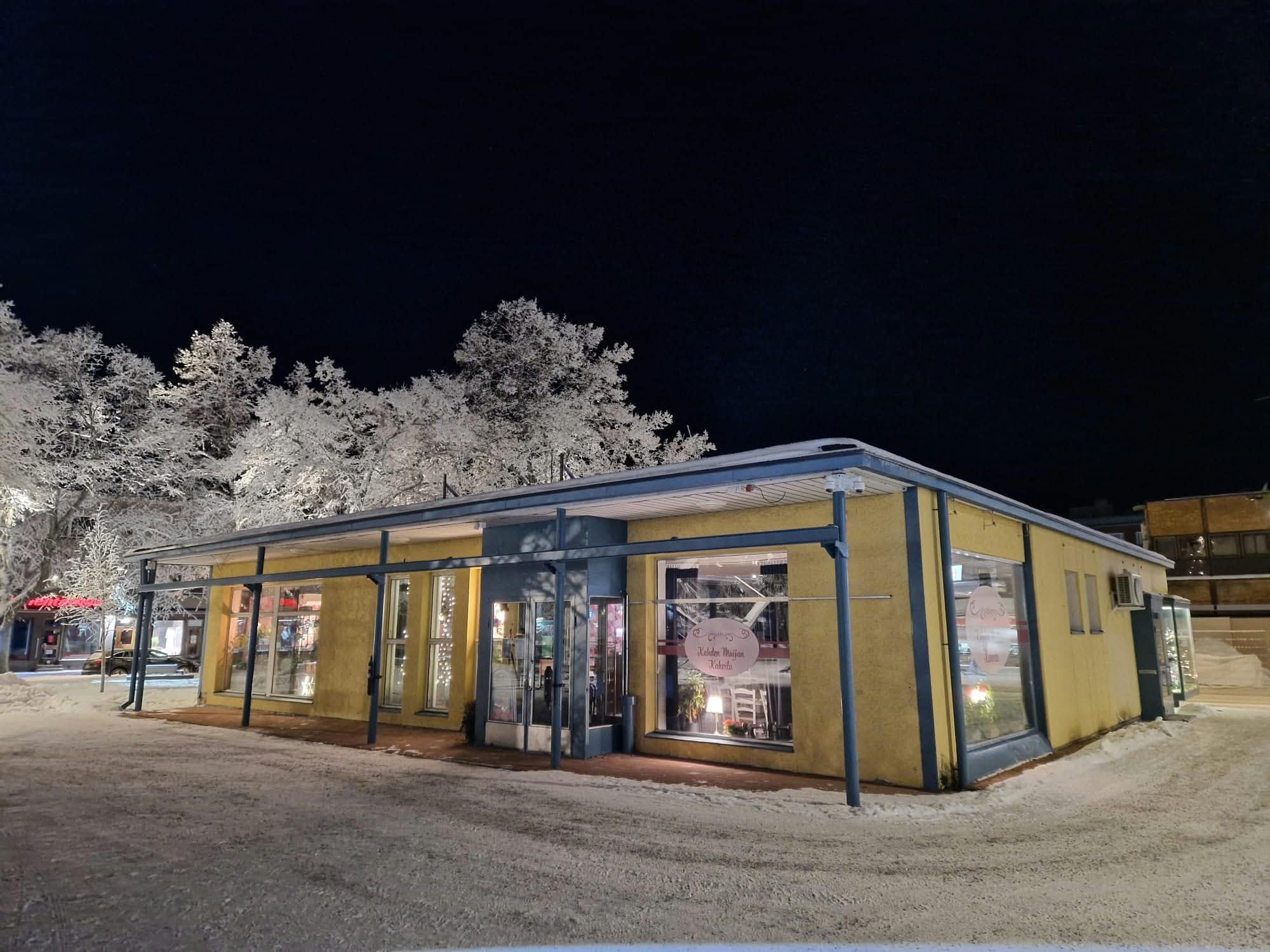 Kahvilan keltainen julkisivu tummana talviyönä — ikkunoista hehkuu lämmin valo ja lumipeitteiset logotarrat kertovat, että kahvila on auki.