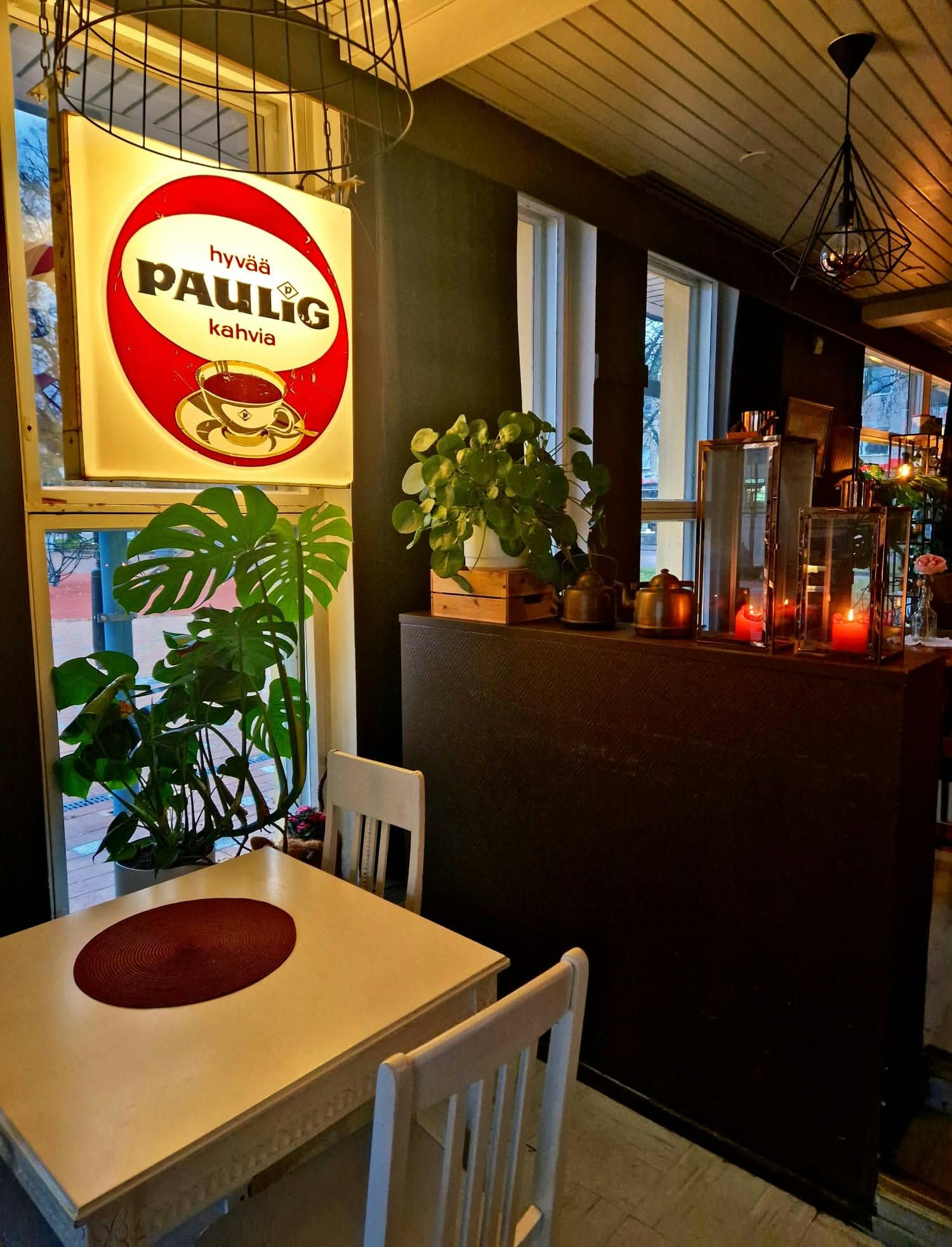 Pieni valkoinen pöytä ja tuolit ikkunan ääressä, seinällä hehkuva Paulig-mainos, palavat kynttilät ja vehreä kasvi.